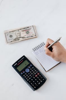 Close-up of hand writing in a notepad with a calculator and dollar bill on a desk, focusing on budgeting.